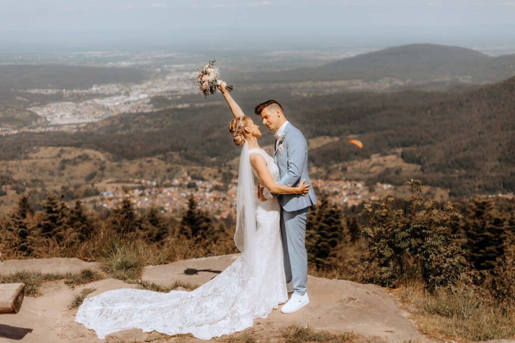 Schönes Brautpaar mit Brautstrauß trifft sich zum First Look an der Teufelsmühle in Loffenau mit schöner Aussicht. Im Hintergrund ist der Nordschwarzwald mit einem Gleitschirmfliegen.