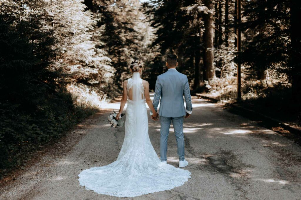 Schönes Brautpaar mit Schleppe und Brautstrauß von hinten im Wald. Bräutigam trägt Anzug in hellblau.