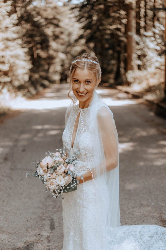 Braut im engen weißen Kleid, Brautstrauß und Haarteil im Wald.