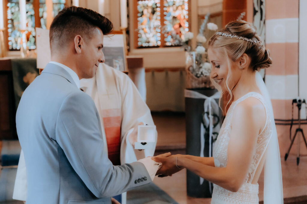 Braut zieht in der Kirche dem Bräutigam einen Ringer an den Finger. Der Pfarrer kündigt den Ringtausch an.