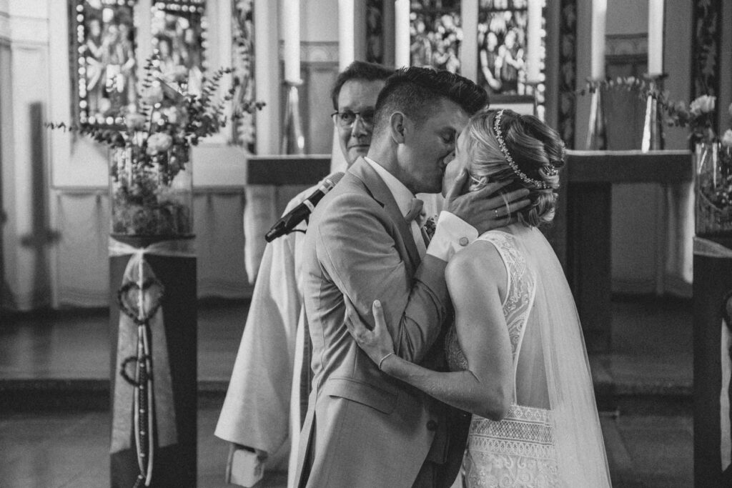 Braut und Bräutigam küssen sich am Alter vor dem Pfarrer. Schwarz weiß Foto.