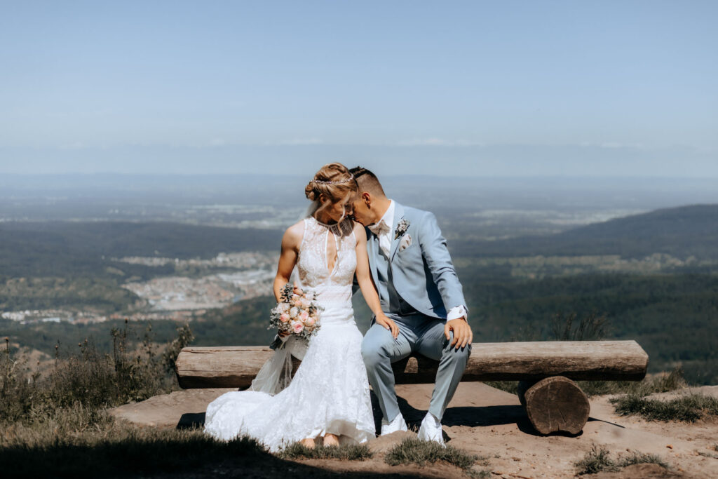 Schönes Brautpaar mit Brautstrauß sitzt auf einer Bank an der Teufelsmühle in Loffenau mit schöner Aussicht. Im Hintergrund ist der Nordschwarzwald.