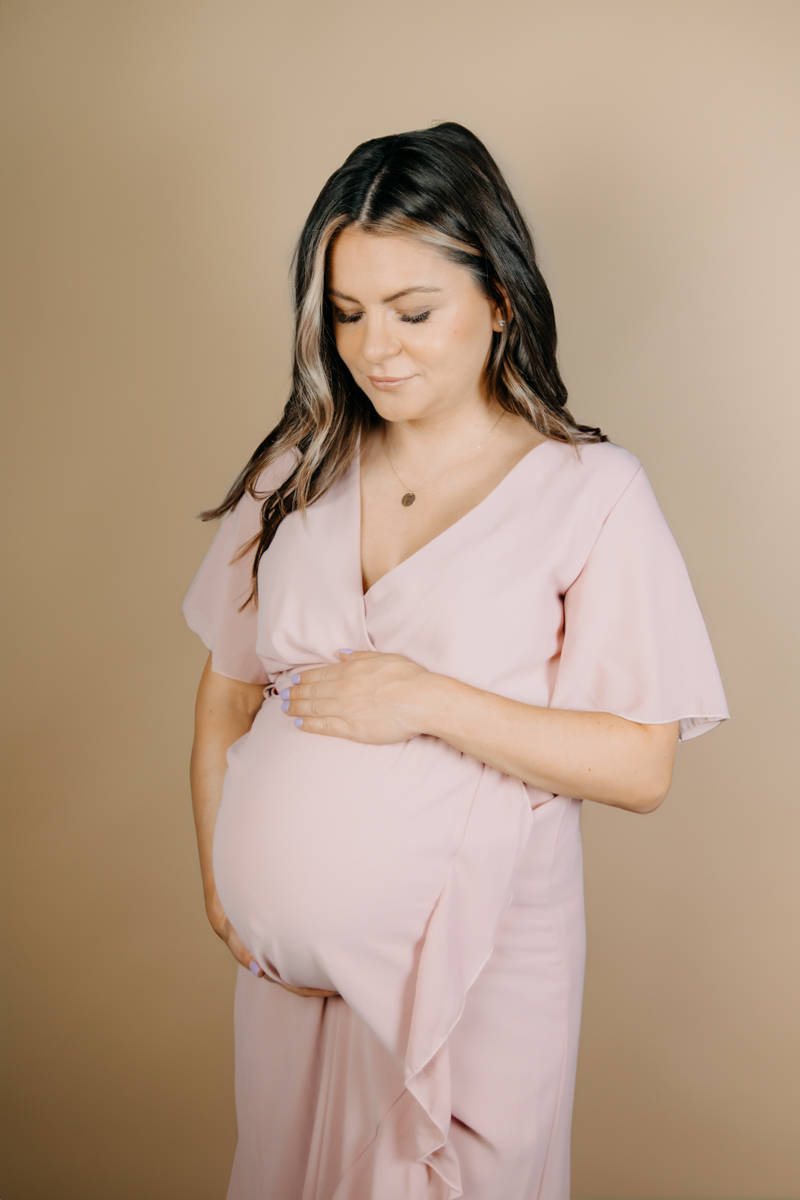 Schwangere Frau beim Babybauchshooting in rosafarbenem Kleid vor Leinwand.